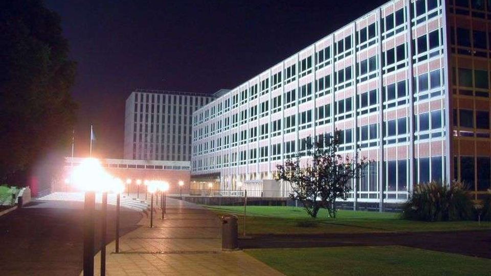 Veduta notturna di un complesso di edifici moderni a più piani con facciate a griglia di finestre illuminate. In primo piano un ampio vialetto pavimentato con lampioni, affiancato da prati e alcuni alberi.