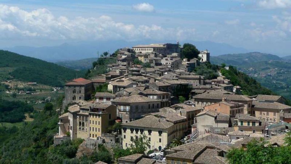 Veduta panoramica di un borgo su una collina, con case addossate tra loro e tetti in tegole. Sullo sfondo si vedono colline e montagne sotto un cielo azzurro con nuvole sparse.