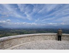 Terrazza panoramica in pietra con parapetto semicircolare e pavimentazione in ciottoli, affacciata su una vasta valle verde con campi e centri abitati. Sullo sfondo si vedono montagne lontane sotto un ampio cielo azzurro con nuvole sparse.