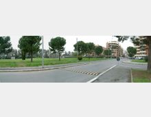 Strada urbana con rotatoria e attraversamento rialzato, affiancata da un’ampia area verde con pini. Sullo sfondo si vedono edifici residenziali multipiano e lampioni lungo la carreggiata.