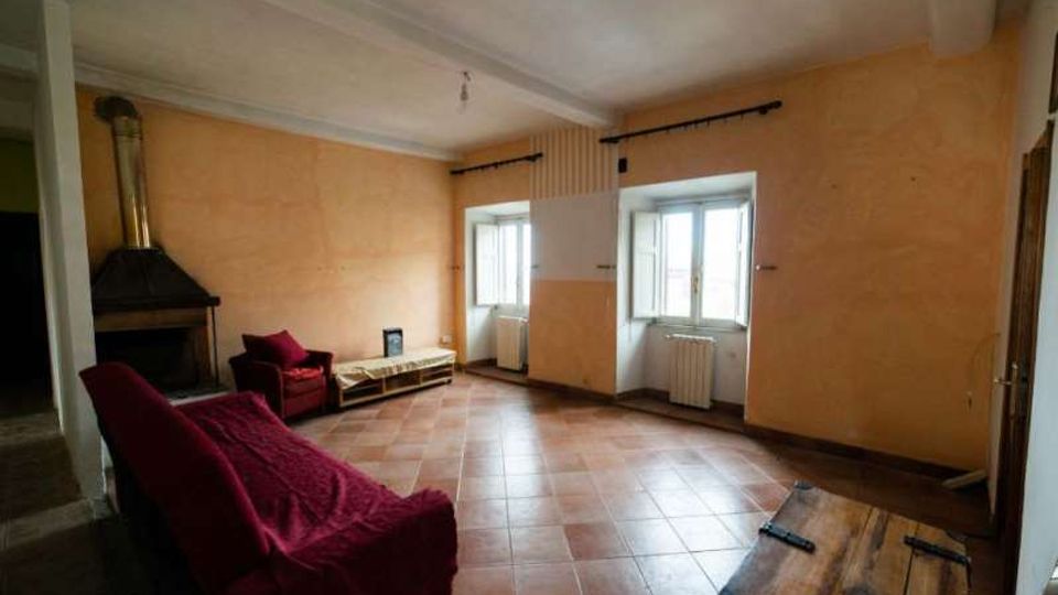 Ampio soggiorno con pareti color pesca, pavimento in piastrelle di terracotta e soffitto bianco con travi. Sono visibili un camino ad angolo, divani rossi, un tavolino in legno e due finestre incassate con radiatori sottostanti.