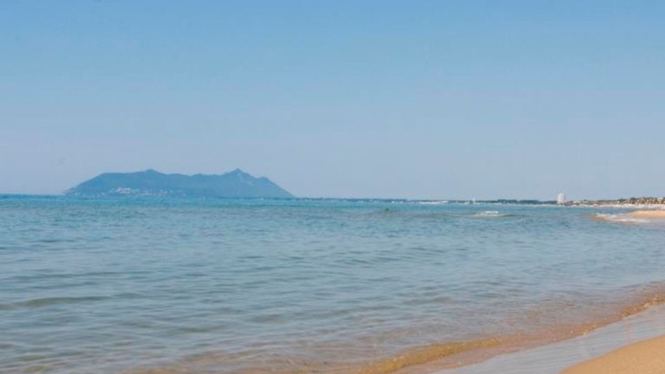 Spiaggia sabbiosa con battigia in primo piano e mare calmo e trasparente che si estende verso l’orizzonte. In lontananza si vede una costa bassa e una sagoma montuosa sotto un cielo azzurro limpido.
