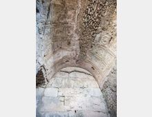 Soffitto a volta in pietra di un ambiente antico, con pannelli decorativi a cassettoni e rosoni scolpiti. Parte della volta è deteriorata, con intonaco mancante e pietrame a vista sopra un arco in muratura.