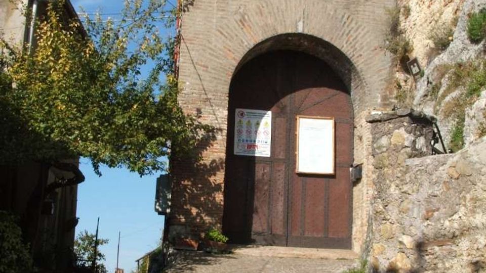 Porta d’ingresso in mattoni di un borgo medievale, con grande arco a tutto sesto e portone in legno lungo una strada acciottolata in salita. Ai lati si vedono mura in pietra, vegetazione e cavi aerei sotto un cielo sereno.