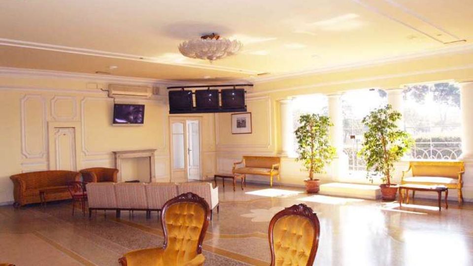 Ampia hall interna con soffitto chiaro, lampadario centrale e pareti con modanature, arredata con poltrone e divani in stile classico e tavolini. Grandi finestre illuminano la sala e affacciano su un balcone, con piante in vaso vicino ai serramenti e schermi TV montati sulla parete.