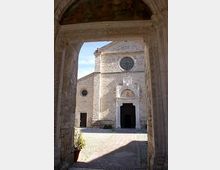 Vista attraverso un arco in pietra di una chiesa in muratura chiara con facciata semplice, rosone circolare e portale centrale con timpano. Davanti all’ingresso si apre un piccolo sagrato lastricato illuminato dal sole.