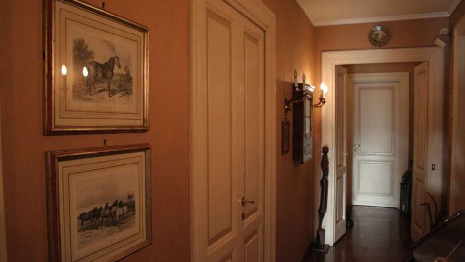 Corridoio interno di una casa con pareti color ocra, pavimento scuro e porte bianche a pannelli lungo il passaggio. Alle pareti sono appesi quadri incorniciati e una lampada a parete illumina l’ingresso verso una porta in fondo.
