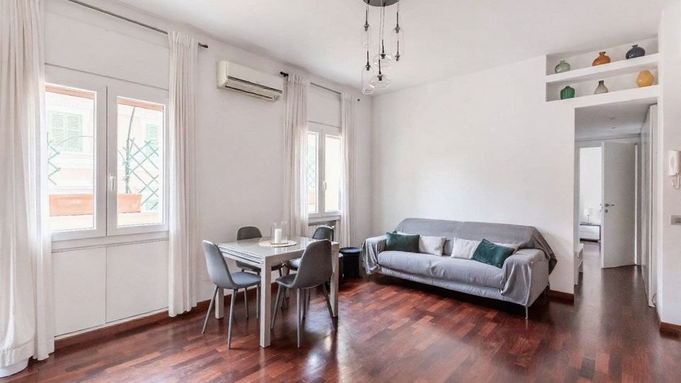 Soggiorno luminoso con pavimento in parquet scuro, pareti bianche e grandi finestre con tende chiare. Nella stanza ci sono un tavolo da pranzo con quattro sedie, un divano grigio e una lampada a sospensione, con passaggio verso altre stanze e mensole con vasi decorativi.