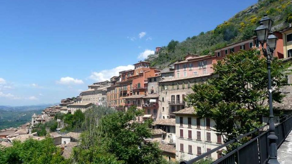 Vista panoramica di un borgo collinare con edifici in pietra e intonaco dai colori caldi disposti in terrazze lungo il pendio. In primo piano si vede una passeggiata con ringhiera e lampioni, circondata da alberi e vegetazione, con valle sullo sfondo sotto un cielo sereno.