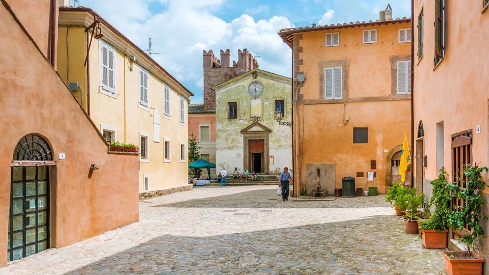 Piazza acciottolata in un borgo storico, circondata da edifici dai colori pastello con persiane e facciate in intonaco. Sullo sfondo si vede una piccola chiesa con portale centrale, orologio sulla facciata e campanile in pietra.