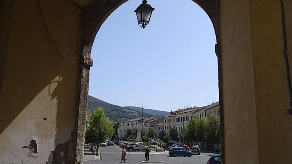 Passaggio ad arco coperto con pareti intonacate e lampione sospeso, che incornicia l’uscita su una piazza pavimentata in pietra. Sullo sfondo si vedono edifici in fila lungo la strada, auto parcheggiate, alberi e colline sotto un cielo sereno.