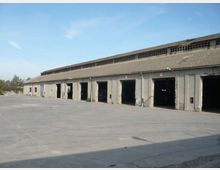 Lungo capannone industriale in cemento con tetto a falda e una fila di ampie aperture tipo portoni, affacciato su un grande piazzale asfaltato. Sul tetto corre una fascia di finestre alte e strette, con cielo sereno sullo sfondo.