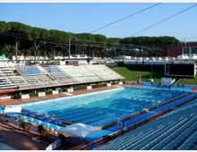 Piscina olimpionica all’aperto con corsie delimitate, circondata da grandi tribune in cemento e sedute. Sullo sfondo si vedono cavi e strutture di copertura, un grande schermo e una fascia di alberi.