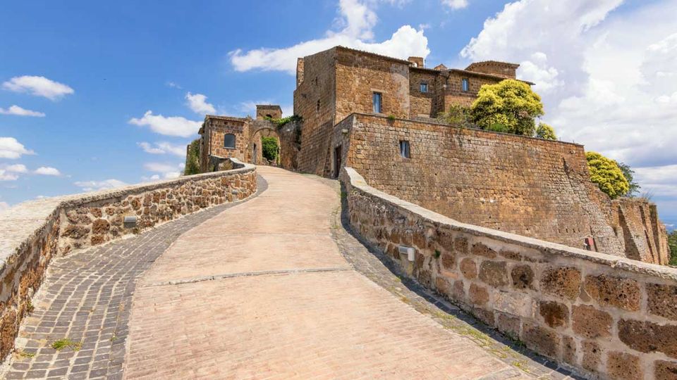 Rampa in pietra e mattoni con muretti laterali che conduce all’ingresso ad arco di un castello in pietra con alte mura e edificio fortificato. Sullo sfondo cielo azzurro con nuvole e vegetazione affacciata sulle mura.