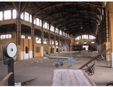 Interno di un grande capannone industriale dismesso, con soffitto alto a capriate metalliche e lunghe file di finestre che illuminano una navata centrale. Pavimento in cemento con detriti sparsi e alcuni carrelli e attrezzature abbandonate in primo piano.