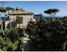 Villa storica in pietra con tetto in tegole e piccola cupola, circondata da un giardino mediterraneo con palme e pini marittimi. Un viale rettilineo conduce all’edificio, con il mare visibile sullo sfondo sotto un cielo sereno.