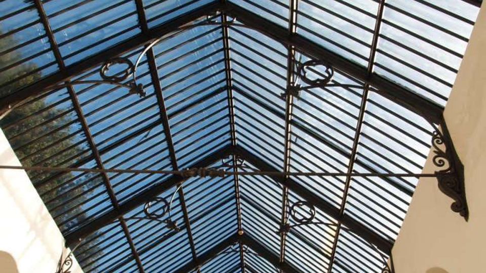 Interno di una galleria coperta con tetto a falde in vetro sorretto da una struttura metallica scura con elementi decorativi. Lungo i lati si vedono pareti chiare con archi e grandi finestre, illuminate dalla luce naturale che filtra dal lucernario.