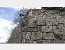 Muro di fortificazione in grandi blocchi di pietra con angolo sporgente, sormontato da una struttura metallica simile a una lanterna o posto di guardia. Su un lato è visibile una targa in pietra con l’iscrizione “Via Gregoriana”, con alberi spogli e cielo sullo sfondo.