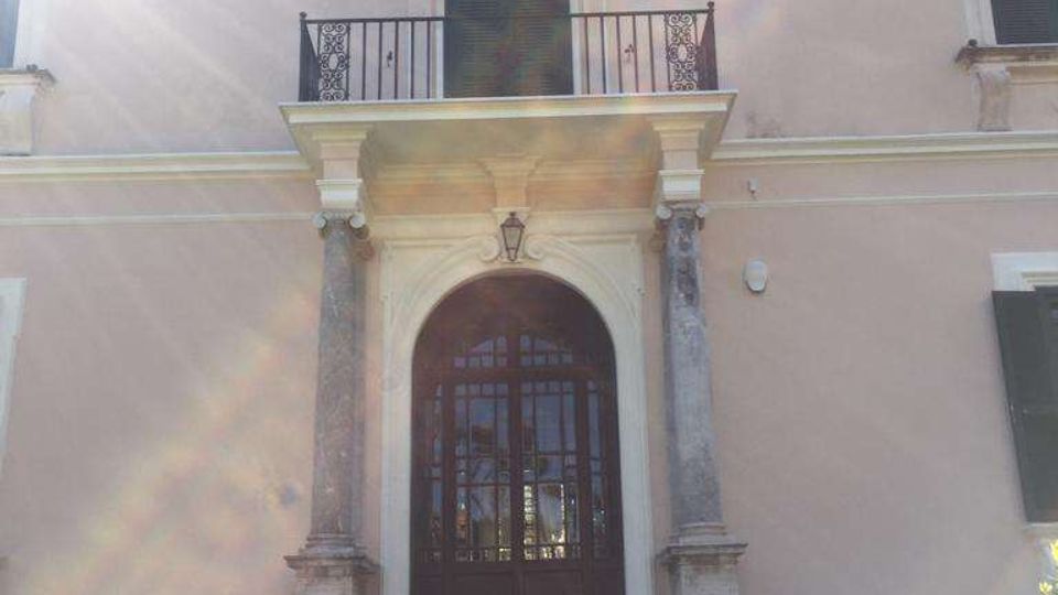 Facciata di una villa storica intonacata color crema, con scalinata in pietra che conduce a un portale ad arco incorniciato da colonne. Sopra l’ingresso c’è un balcone con ringhiera in ferro battuto e finestre con persiane scure.