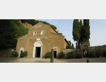 Facciata di una chiesa romanica in pietra con portale ad arco e piccole finestre, affacciata su un ampio piazzale sterrato con vasi di piante. A destra si vede un muro in pietra con ingresso ad arco e alti cipressi, sullo sfondo una collina alberata.