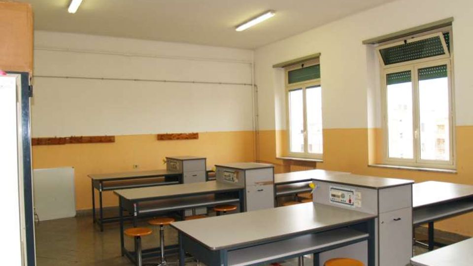 Aula laboratorio con banchi di lavoro in metallo e sgabelli, pavimento in cemento e pareti bianche con fascia gialla. Due finestre con avvolgibili e lampade fluorescenti a soffitto illuminano lo spazio.