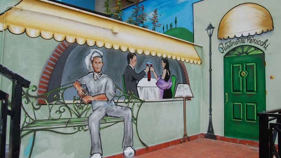 Murale su una parete esterna che raffigura una scena di ristorante con panchina, cameriere e tavolo apparecchiato sotto una tenda dipinta. Accanto c’è una porta verde con insegna “Ristorante Pericchi”, una lampada a muro e pavimentazione in piastrelle di terracotta.
