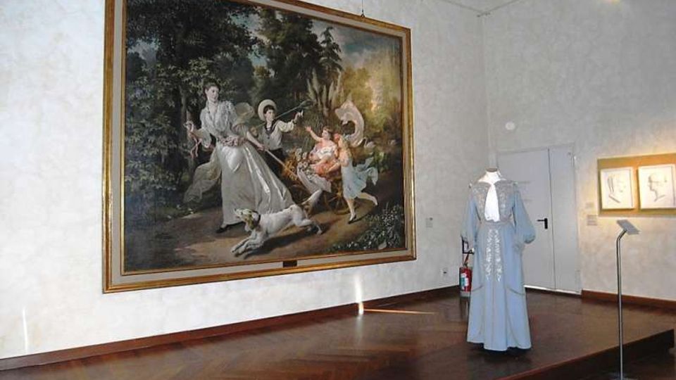 Sala museale con pavimento in parquet e pareti bianche, dominata da un grande dipinto incorniciato appeso alla parete. Al centro della stanza è esposto su una pedana un abito storico, con faretti e alcune opere incorniciate visibili sulla parete laterale.