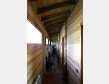 Corridoio interno stretto con pareti e soffitto in legno a vista, travi scure inclinate e pavimento piastrellato. Sul lato sinistro ci sono finestre che illuminano l’ambiente, mentre a destra si aprono porte lungo il corridoio fino a una porta in fondo.