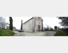 Piccola chiesa in pietra con facciata a timpano e portale centrale, situata su uno spiazzo di ghiaia. Intorno si vedono cipressi e altri alberi, con una strada e un muretto lungo il bordo dell’area.