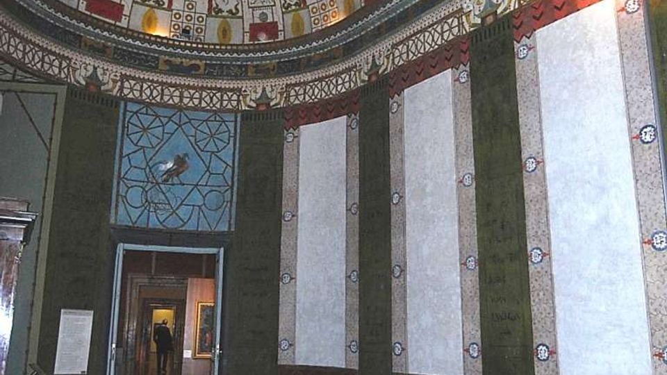 Interno di una sala circolare con pareti scandite da pannelli verticali e zoccolatura decorata, pavimento in legno e alcune sedie lungo la parete. Il soffitto a cupola è riccamente dipinto con motivi ornamentali e figure, con un’apertura che conduce a un corridoio di stanze.