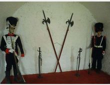 Interno di una piccola stanza con pareti bianche grezze e pavimento rosso, allestita come esposizione militare. Al centro sono appese due alabarde incrociate e ai lati sono disposti fucili e altre armi su supporti.