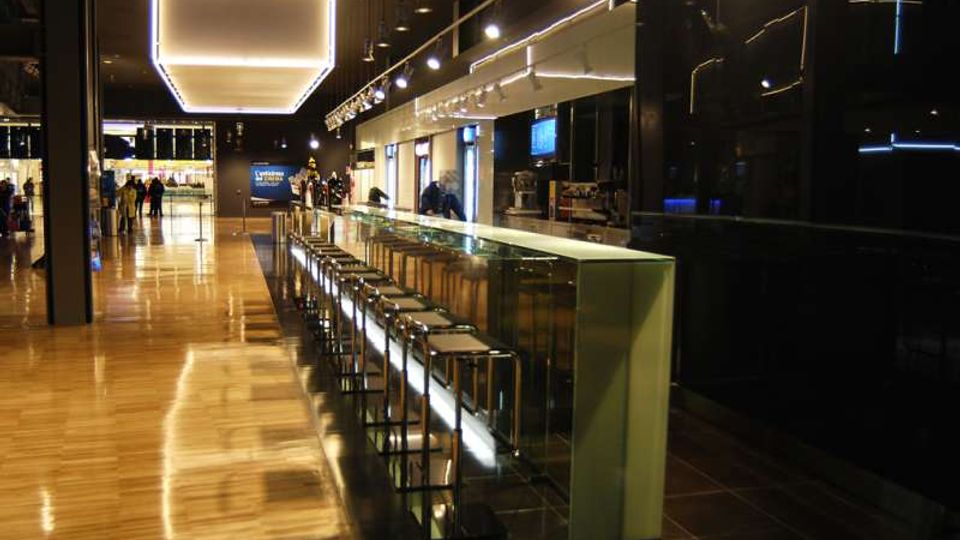 Interno di un moderno bar in stile minimalista, con lungo bancone vetrato e fila di sgabelli in metallo su pavimento lucido. Soffitto con grandi pannelli luminosi e luci a sospensione, pareti scure e area di servizio sullo sfondo.
