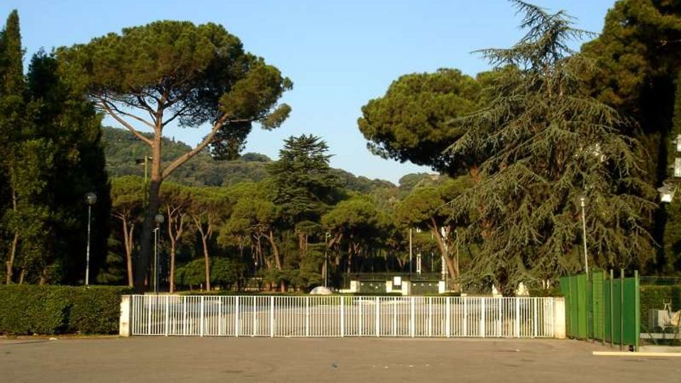 Ampio piazzale asfaltato con una lunga cancellata bianca sullo sfondo, circondato da una fitta area verde con pini ad ombrello e altri alberi alti. Sul lato destro si vede una recinzione verde e alcuni lampioni lungo il perimetro.