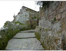 Sentiero in pietra a gradoni che sale tra grandi rocce e una parete rocciosa, con muschio e licheni sulle superfici. In alto si vede un muretto in muratura con ringhiera metallica lungo il bordo.
