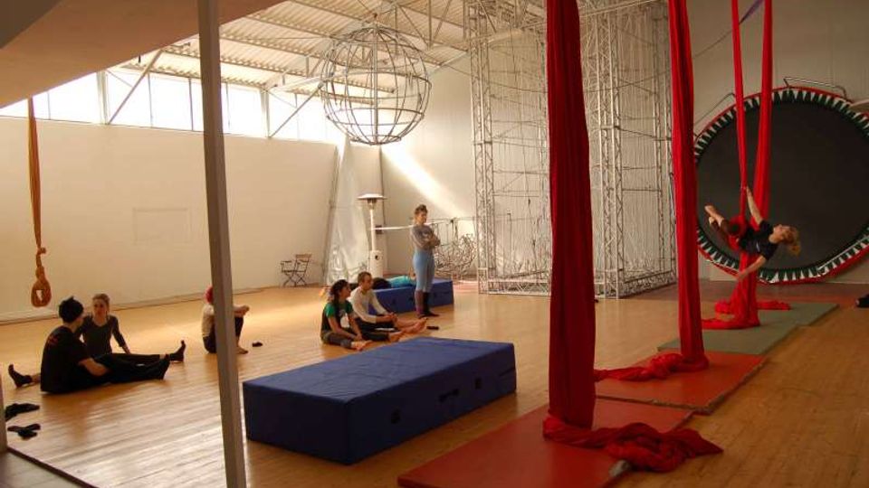 Ampio studio per acrobatica aerea con pavimento in legno e grandi finestre alte, illuminato da luce naturale. Nella sala si vedono tessuti aerei rossi appesi al soffitto, materassi e tappeti di sicurezza, una struttura metallica e un grande trampolino circolare sul lato destro.