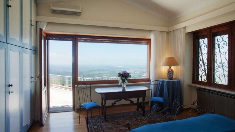 Camera da letto con pavimento in legno, armadio azzurro e letto con copriletto blu, affacciata su una grande porta finestra scorrevole che conduce a un terrazzo con vista panoramica. Nella stanza ci sono una scrivania con due sedie, tende bianche e una finestra laterale con inferriate decorative.