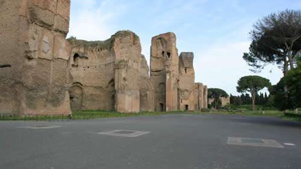 Rovine di antiche mura in mattoni e pietra con grandi arcate e torri parzialmente crollate, lungo un ampio viale asfaltato. Sullo sfondo si vedono aree verdi con pini e cielo aperto.