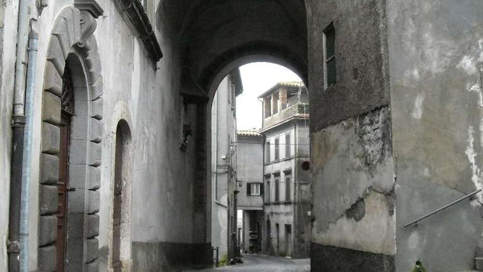 Vicolo stretto con pavimentazione in pietra che curva sotto un grande arco in muratura collegato a edifici storici. Sui lati si vedono facciate intonacate e consumate, portali in pietra, finestre e alcuni gradini che salgono lungo il muro destro.