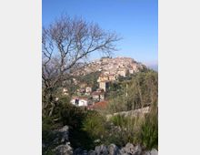 Veduta panoramica di un borgo collinare con case in pietra e tetti in coppi, disposto a gradoni sul pendio. In primo piano ci sono rocce, erba e rami di alberi spogli, con vegetazione e cielo sereno sullo sfondo.