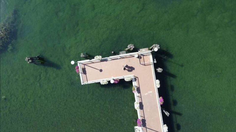 Vista dall’alto di un molo a forma di “L” che si estende su un lago verde, con ringhiere e fioriere lungo i bordi e alcune piccole strutture alle estremità. L’acqua circonda completamente la passerella, con ombre proiettate sul fondale.