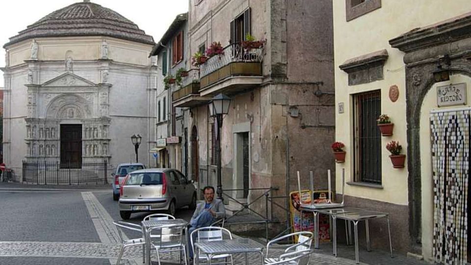 Piccola piazza con pavimentazione in pietra e tavolini metallici all’aperto davanti a edifici storici con balconi e fioriere. Sullo sfondo si vede una chiesa in pietra chiara con facciata decorata e tetto ottagonale, con auto parcheggiate lungo la strada.