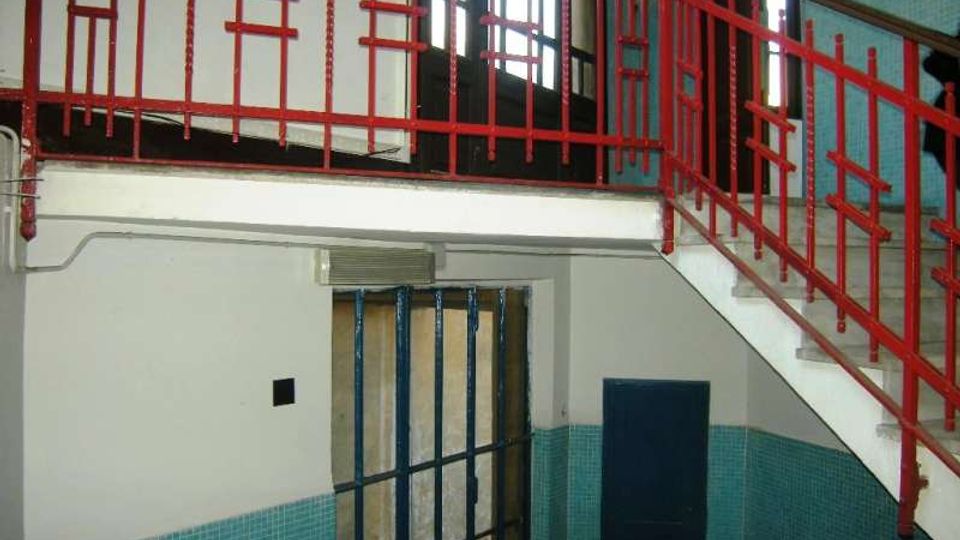 Interno di un edificio con scala e pianerottolo, caratterizzato da una ringhiera metallica rossa geometrica e pareti rivestite a metà con piastrelle turchesi. In basso si vede un corridoio con una porta chiusa e un’apertura con inferriata metallica, illuminata da finestre alte sul fondo.