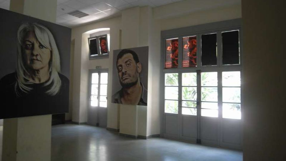 Interno di una galleria o sala espositiva con pavimento lucido e soffitto a pannelli, illuminata da grandi porte-finestre a vetri e finestre alte. Su pilastri e pareti sono appesi grandi quadri con ritratti, disposti lungo lo spazio aperto.