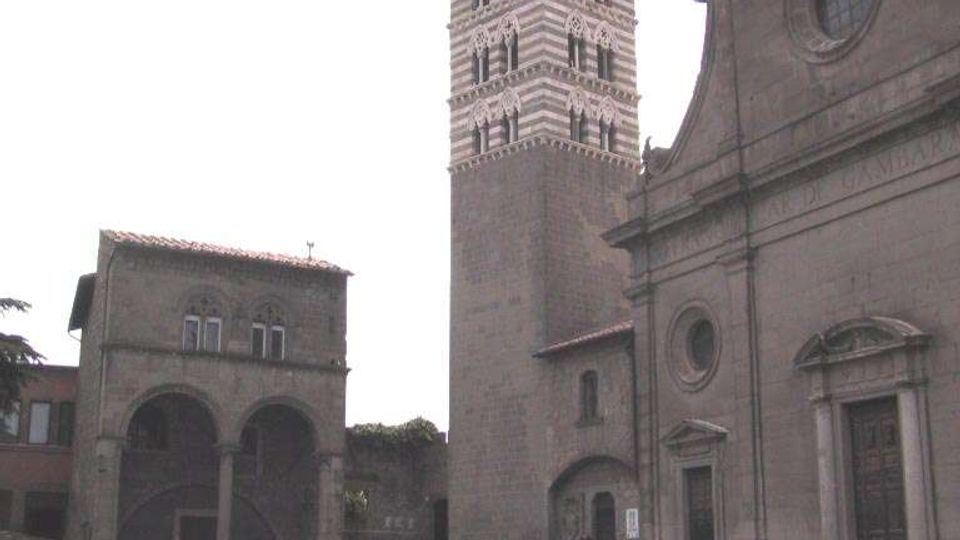 Piazza lastricata con una chiesa in pietra e ampia scalinata d’ingresso, affiancata da un alto campanile a fasce orizzontali e da un edificio con portico ad archi. Sullo sfondo si vedono altri edifici storici che delimitano lo spazio urbano.