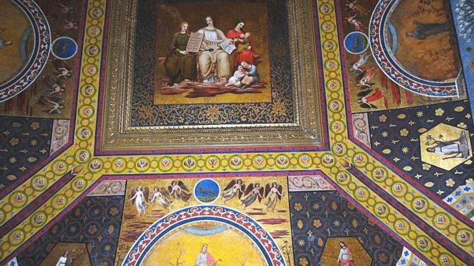 Soffitto decorato di un interno storico con riquadri dipinti e cornici dorate, con un grande pannello centrale figurativo e motivi ornamentali geometrici. Intorno si vedono medaglioni e pannelli con scene e figure su fondo blu e oro, tipici di una volta affrescata.