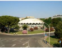 Edificio moderno a cupola bianca con struttura reticolare visibile alla base, circondato da alberi e aiuole fiorite. In primo piano si vede una grande rotatoria stradale con corsie e segnaletica, sullo sfondo altri edifici urbani.