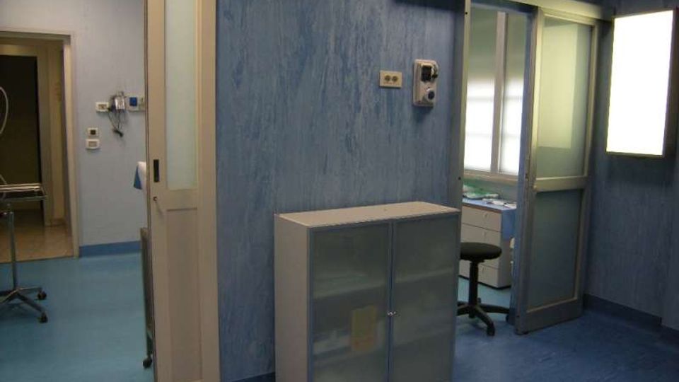Interno di una stanza clinica con pareti e pavimento blu, porte scorrevoli con pannelli in vetro e una finestra luminosa. Sono presenti un armadietto basso, attrezzature a parete e un’area adiacente con carrello metallico e postazione con sgabello.