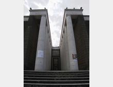 Facciata monumentale di un edificio pubblico con due alte file simmetriche di colonne cilindriche in pietra chiara, affiancate da pareti scure e raggiunte da un’ampia scalinata. Al centro si vede un ingresso incassato con porta vetrata, sotto un cielo nuvoloso.