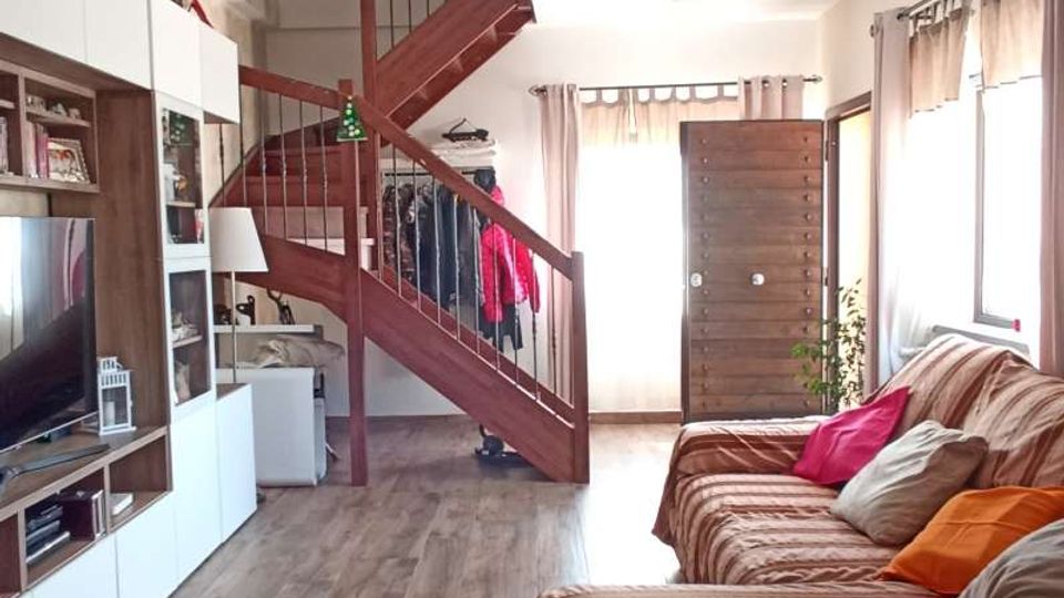 Soggiorno luminoso con pavimento in legno, grande divano a righe lungo la parete e parete attrezzata con TV e scaffali. Sul fondo si vede una scala interna in legno con ringhiera e una porta d’ingresso, con finestre laterali dotate di tende chiare.