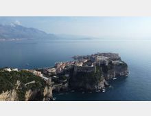 Veduta panoramica di una cittadina costiera con edifici storici e fortificazioni su un promontorio roccioso a picco sul mare. In lontananza si vedono montagne e una costa che si estende lungo l’orizzonte, con acque calme intorno al promontorio.